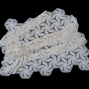 May include: Cream-colored, intricately crocheted doily or tablecloth with a repeating geometric pattern. The edges are scalloped, and the piece is displayed against a black background. The design features spiraling shapes.