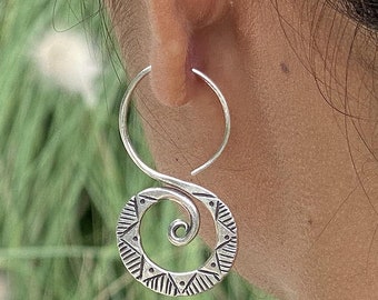 May include: Silver hoop earrings with a spiral design and geometric patterns. The earrings are shown on a person's ear.