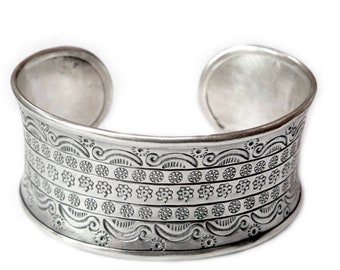 May include: A silver cuff bracelet with an intricate embossed design featuring repeating patterns of swirls, circles, and flowers.