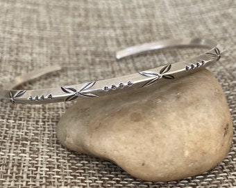 May include: A silver bangle bracelet with a delicate floral design. The bracelet is open and has a simple, elegant style.
