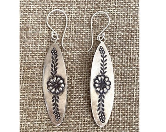 May include: A pair of silver dangle earrings with a floral design. The earrings are oval shaped and have a black floral pattern with leaves.