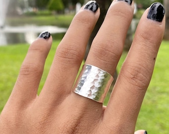 May include: A wide, silver ring with a hammered texture. The ring is a solid band, and the surface has a dimpled appearance. The ring is worn on a finger, with a green background visible.