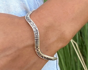 May include: A silver bracelet with a wavy design and intricate etched patterns. The bracelet is worn on a person's wrist.