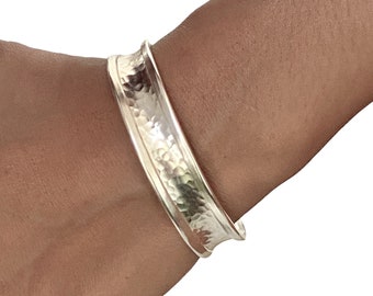 May include: A silver hammered cuff bracelet with a rounded edge. The bracelet is worn on a person's wrist.