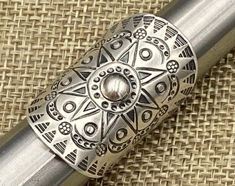May include: A silver ring with a detailed geometric design featuring a central starburst and a circular pattern. The ring is made of metal and has a polished finish.