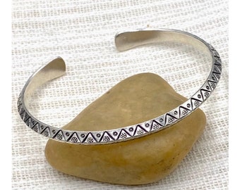 May include: A silver bangle bracelet with a geometric pattern. The bracelet is made of a thin metal and has a simple design.