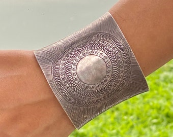 May include: A silver cuff bracelet with an intricate circular design. The bracelet is square-shaped and features a detailed pattern of concentric circles and lines. The metal has a brushed finish, giving it a matte appearance.