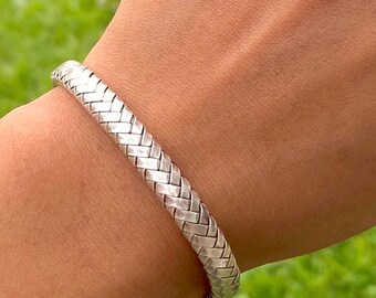 May include: A silver-colored bracelet with a woven design is worn on a person's wrist. The bracelet has a textured surface and a rounded shape. The background includes green grass and a body of water.