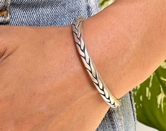 May include: A silver braided cuff bracelet. The bracelet is worn on a person's wrist.