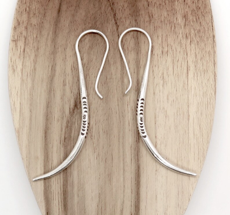 May include: A pair of silver hoop earrings with a curved, textured design. The earrings are made of sterling silver and have a simple, elegant style.