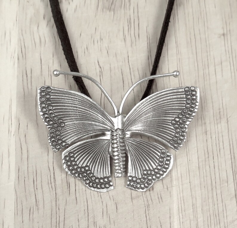 May include: A silver butterfly pendant necklace with a detailed design. The butterfly has a textured surface and is suspended from a black cord.