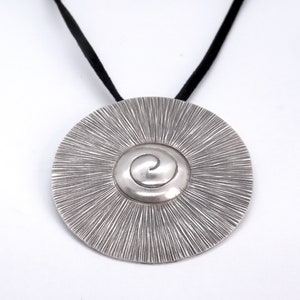 May include: A silver pendant necklace with a circular design featuring a spiral in the centre. The pendant is attached to a black cord.