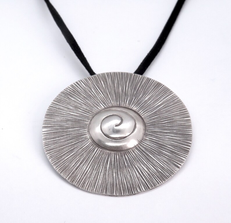May include: A silver pendant necklace with a circular design featuring a spiral in the centre. The pendant is attached to a black cord.