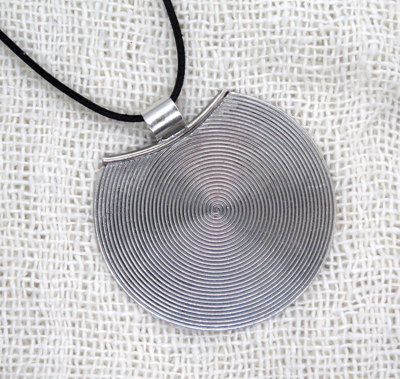 May include: A silver pendant necklace with a circular design featuring concentric circles. The pendant is attached to a black cord.