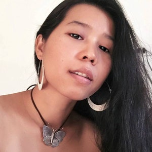 May include: A woman with long black hair is wearing a silver butterfly pendant necklace and silver hoop earrings. The necklace is on a brown leather cord.