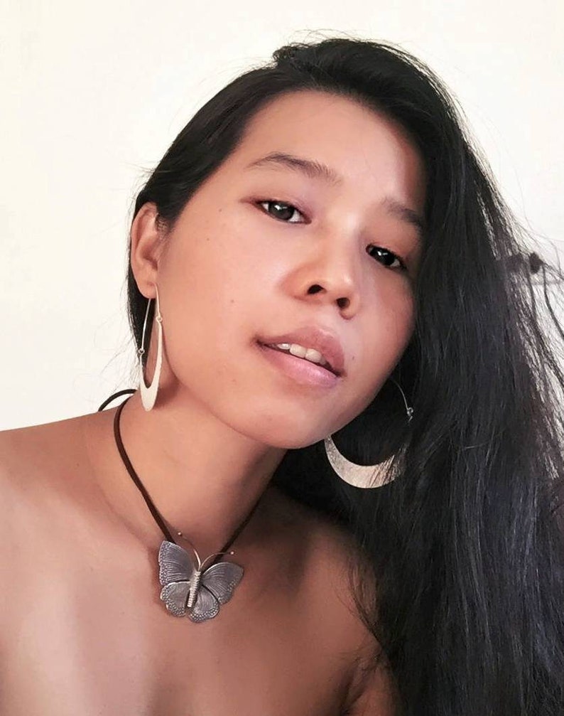 May include: A woman with long black hair is wearing a silver butterfly pendant necklace and silver hoop earrings. The necklace is on a brown leather cord.