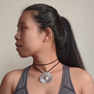May include: A woman wearing a grey tank top with a silver pendant necklace on a brown cord. The pendant is a round, textured disc with a raised centre.