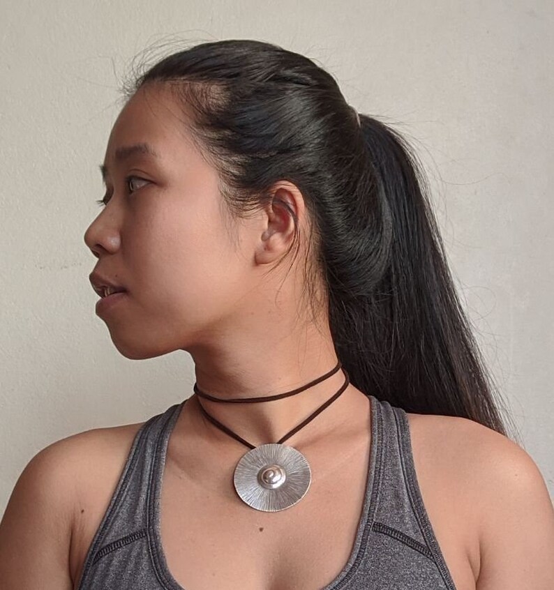 May include: A woman wearing a grey tank top with a silver pendant necklace on a brown cord. The pendant is a round, textured disc with a raised centre.