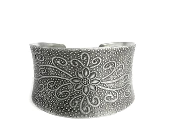 May include: A silver cuff bracelet with a floral design. The bracelet is made of metal and has a textured surface.