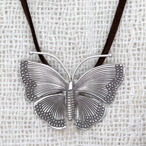 May include: A silver butterfly pendant necklace with a brown suede cord. The butterfly has detailed wings with a textured surface.