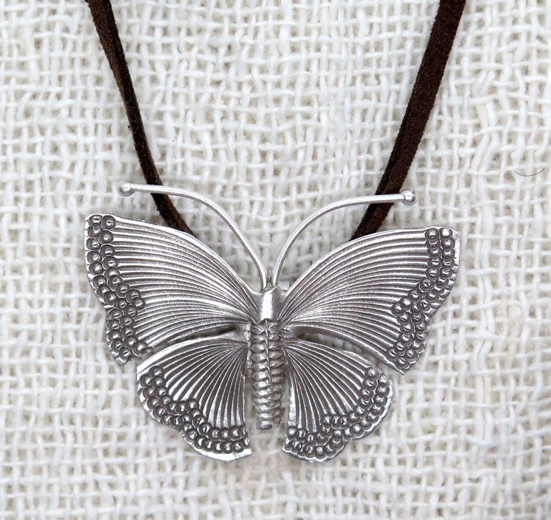 May include: A silver butterfly pendant necklace with a brown suede cord. The butterfly has detailed wings with a textured surface.