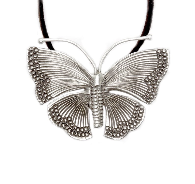 May include: A silver butterfly pendant necklace with a detailed design. The pendant is attached to a black cord.