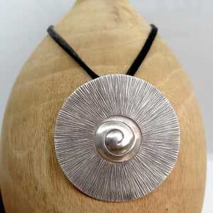 May include: A silver pendant necklace with a circular design featuring a spiral pattern in the centre. The pendant is hanging from a black cord.