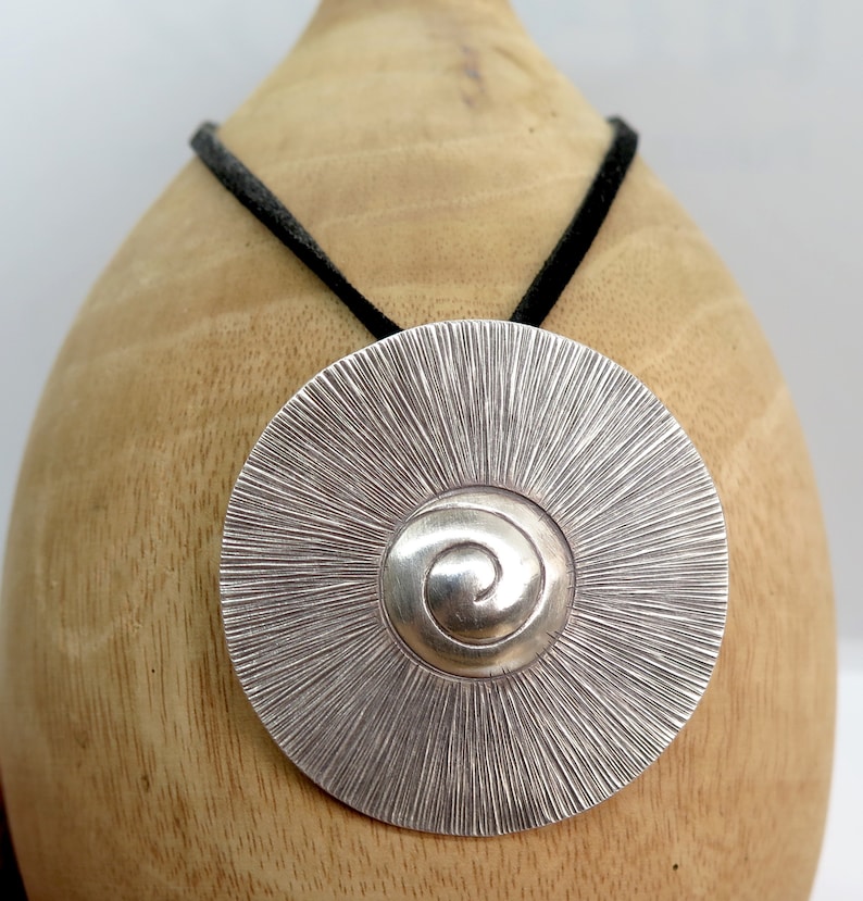 May include: A silver pendant necklace with a circular design featuring a spiral pattern in the centre. The pendant is hanging from a black cord.