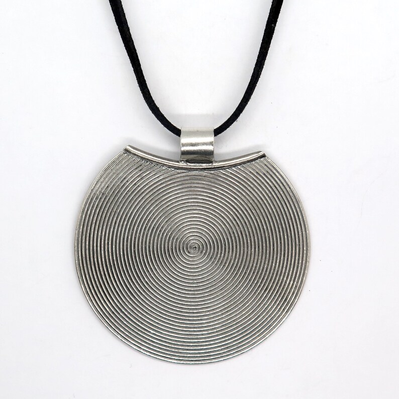 May include: A silver pendant necklace with a circular pendant featuring a concentric circle design. The pendant is attached to a black suede cord.