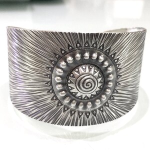 May include: Wide silver cuff bracelet with a sunburst design. The bracelet features a central sun emblem with a spiral center and beaded accents. The textured surface has radiating lines, creating a bold, eye-catching accessory.