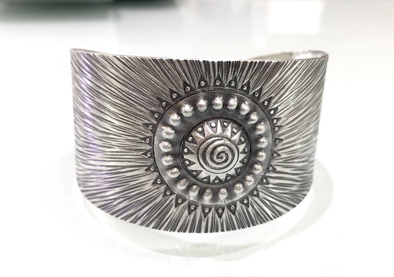 May include: Wide silver cuff bracelet with a sunburst design. The bracelet features a central sun emblem with a spiral center and beaded accents. The textured surface has radiating lines, creating a bold, eye-catching accessory.