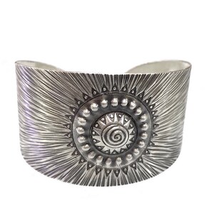 May include: A wide, silver-toned cuff bracelet. The bracelet features a central sunburst design with a spiral center and a textured, radial pattern. The cuff is approximately 2 inches wide.