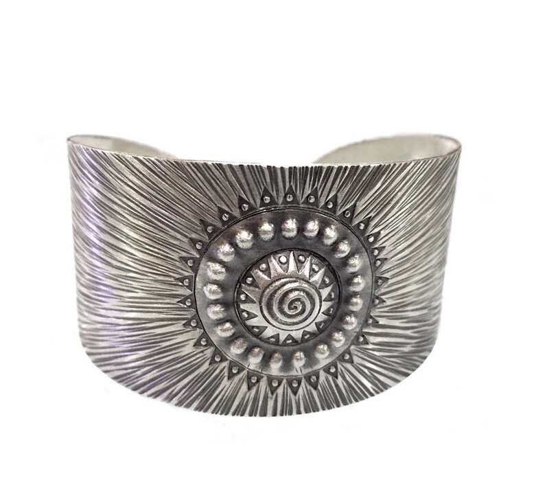 May include: A wide, silver-toned cuff bracelet. The bracelet features a central sunburst design with a spiral center and a textured, radial pattern. The cuff is approximately 2 inches wide.