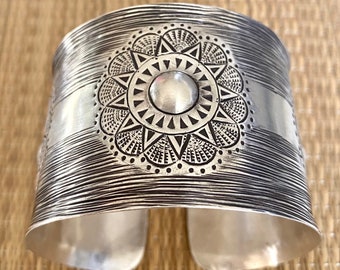 May include: A silver cuff bracelet with a detailed sun design. The sun is surrounded by a circle of smaller circles and the center of the sun is a raised circle.