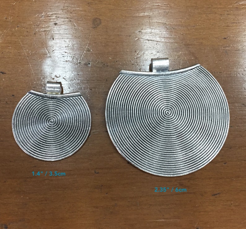 May include: Two silver metal pendants with a spiral design. The smaller pendant measures 1.4 inches or 3.5 cm in diameter. The larger pendant measures 2.35 inches or 6 cm in diameter.