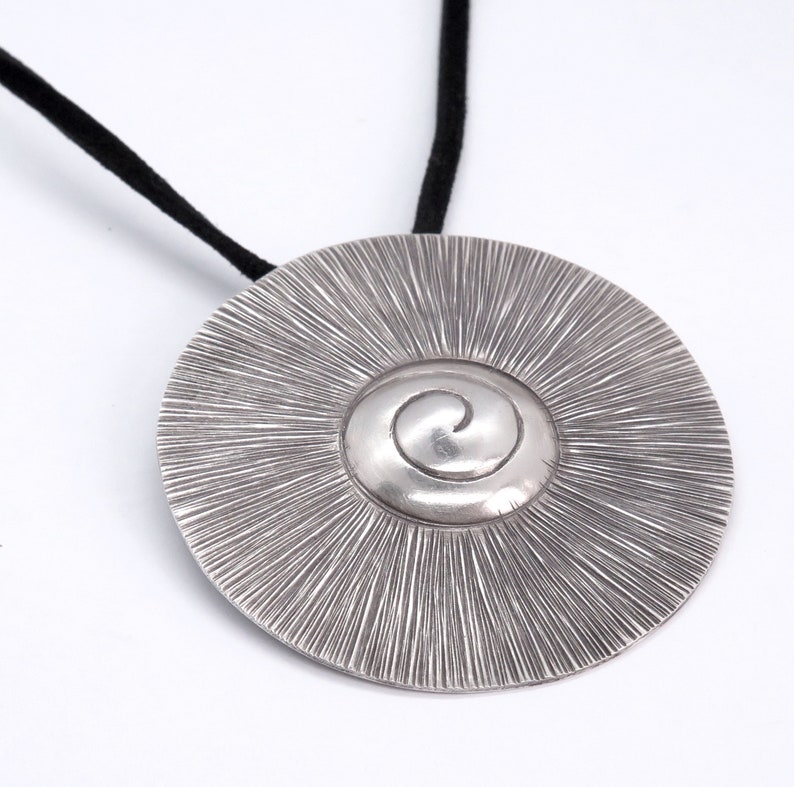 May include: A silver pendant necklace with a circular design featuring a spiral in the centre. The pendant is attached to a black cord.