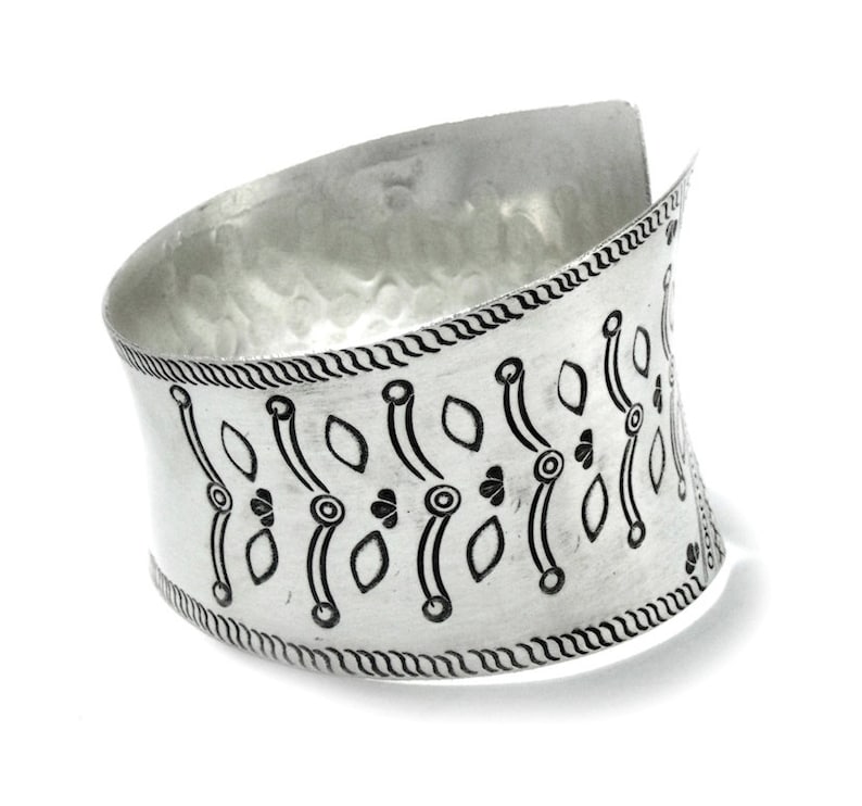 Puede incluir: Pulsera ancha de plata con textura martillada y un patr&oacute;n decorativo negro. La pulsera presenta un dise&ntilde;o curvo y un borde detallado, con un estilo abierto y envolvente.