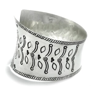 Puede incluir: Un brazalete ancho plateado con textura martillada. El brazalete presenta un patr&oacute;n repetido de dise&ntilde;os de hojas y c&iacute;rculos negros. Los bordes est&aacute;n detallados con un borde trenzado. Esta pieza de joyer&iacute;a est&aacute; dise&ntilde;ada para ser usada en la mu&ntilde;eca.