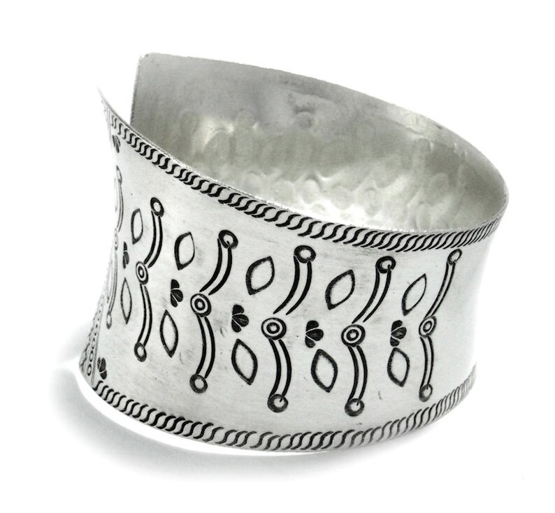 Puede incluir: Un brazalete ancho plateado con textura martillada. El brazalete presenta un patr&oacute;n repetido de dise&ntilde;os de hojas y c&iacute;rculos negros. Los bordes est&aacute;n detallados con un borde trenzado. Esta pieza de joyer&iacute;a est&aacute; dise&ntilde;ada para ser usada en la mu&ntilde;eca.