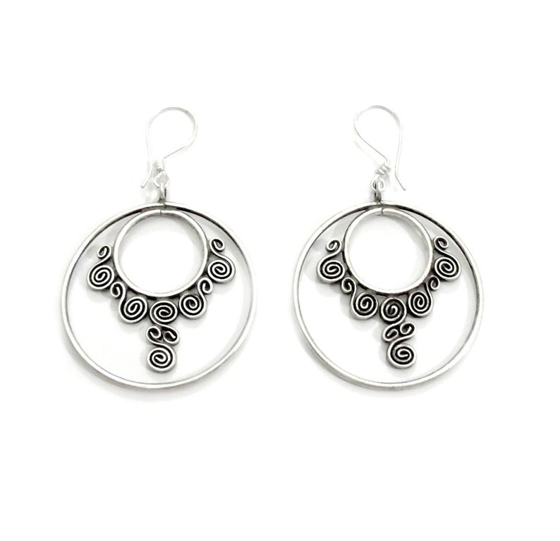 May include: Silver hoop earrings with a delicate, intricate design of swirling circles. The earrings have a double hoop design, with a smaller hoop inside a larger hoop.