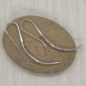 May include: A pair of silver dangle earrings with a curved design and a textured pattern. The earrings are on a light brown stone.
