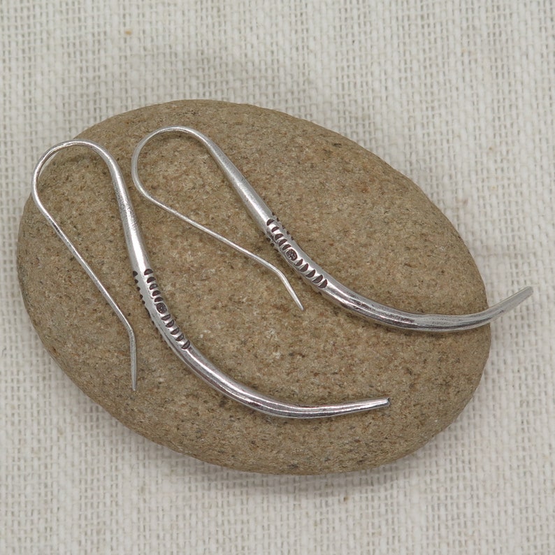 May include: A pair of silver dangle earrings with a curved design and a textured pattern. The earrings are on a light brown stone.