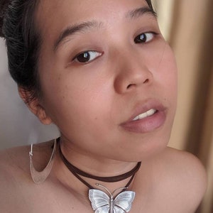 May include: A silver butterfly pendant necklace with a brown leather cord. The pendant is detailed with intricate designs and has a slightly textured finish.