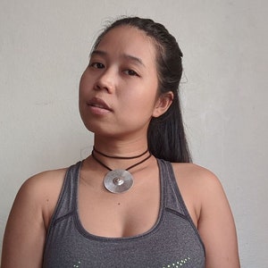 May include: A woman with long dark hair is wearing a grey tank top and a silver pendant necklace with a brown leather cord.