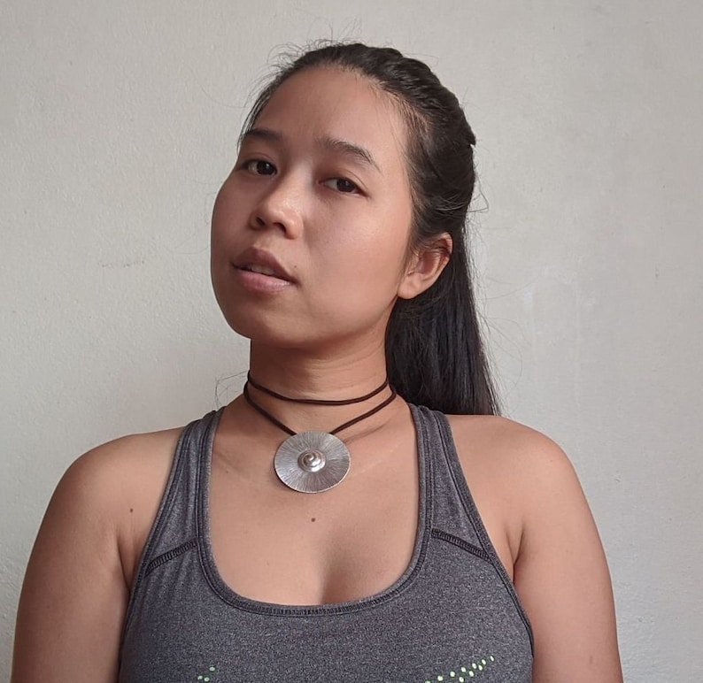 May include: A woman with long dark hair is wearing a grey tank top and a silver pendant necklace with a brown leather cord.
