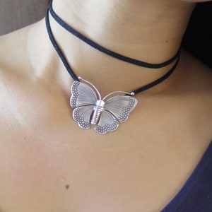May include: A silver butterfly pendant necklace with a black suede choker. The butterfly pendant has a detailed design with a textured surface.