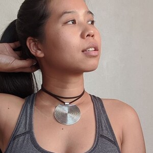 May include: A woman wearing a gray tank top with a silver circular pendant necklace on a brown cord.