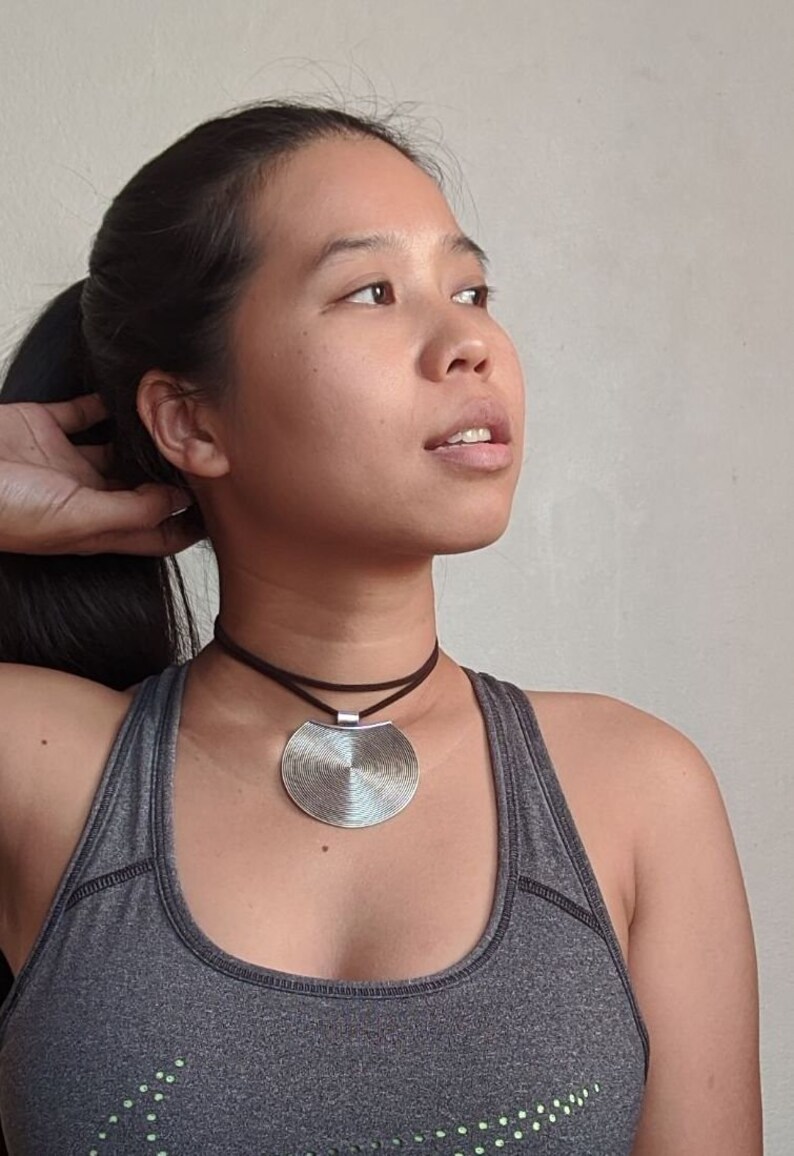 May include: A woman wearing a gray tank top with a silver circular pendant necklace on a brown cord.