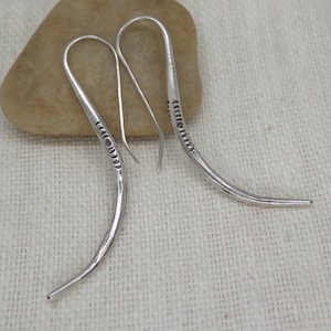 May include: A pair of silver dangle earrings with a curved design and a textured pattern. The earrings are made of metal and have a simple, elegant style.