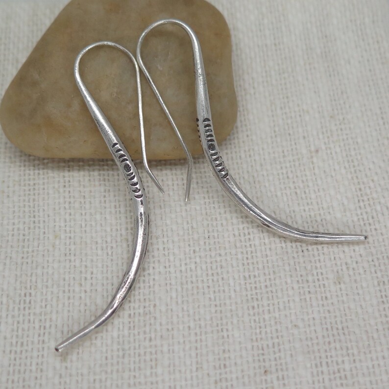 May include: A pair of silver dangle earrings with a curved design and a textured pattern. The earrings are made of metal and have a simple, elegant style.